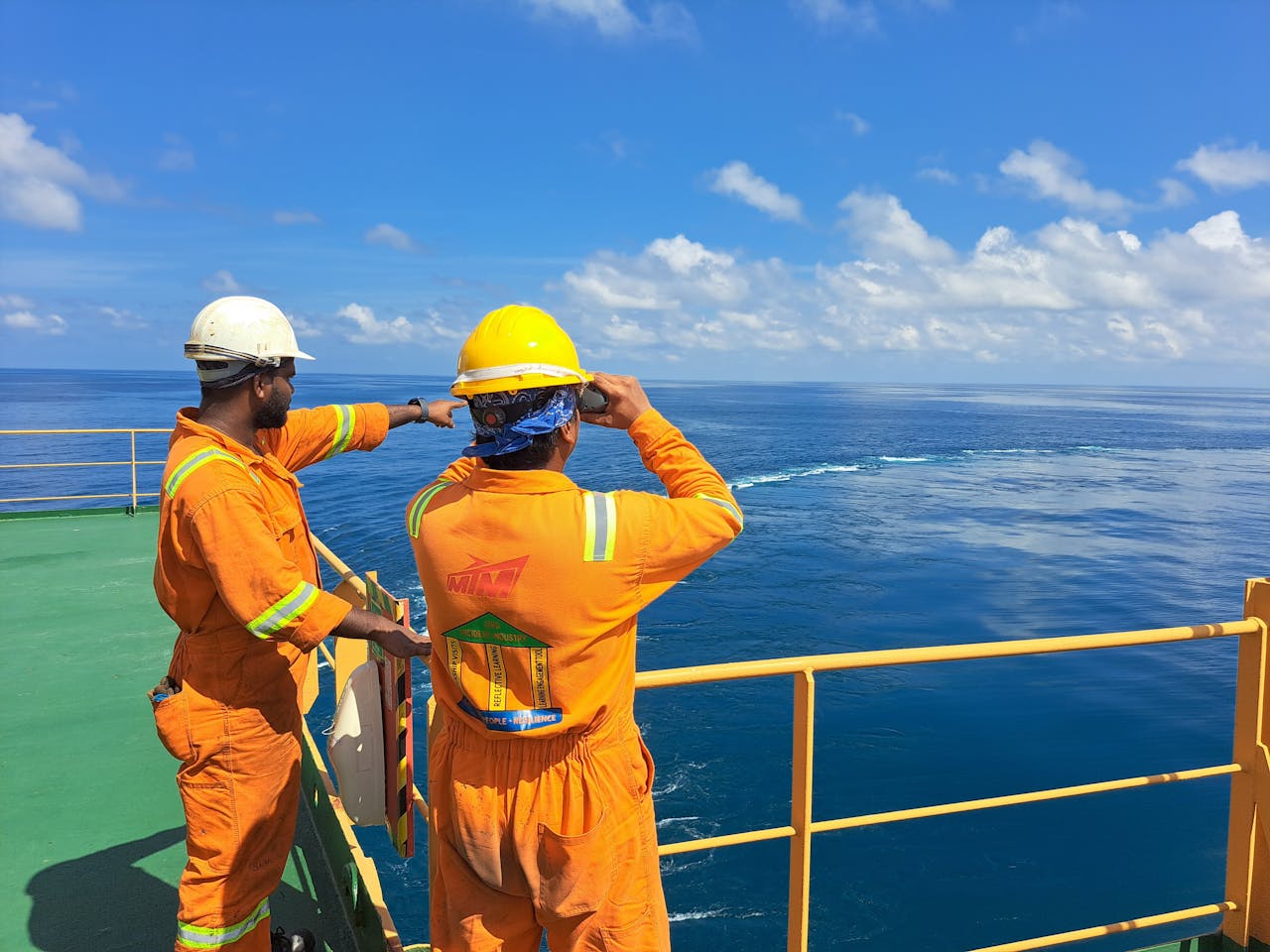contact-img Two workers on an oil rig deck wearing safety gear inspect the ocean horizon under a clear sky.